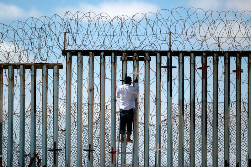 En deporterad mexikansk migrant hänger upp träkors på gränsstängslet för att hedra minnet av migranter som dött på sitt försök till inresa i USA.