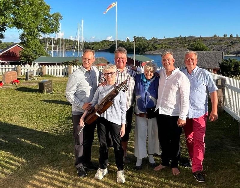 Riko Eklundh (andra från höger) har bjudit in paret Carl och Åsa Unander-Scharin (med nyckelharpa) och sina bröder Fredi och Tom Eklundh (längst till höger) för att delta i konserten Sommarsång i skärgården. I mitten står Monica Aaltonen som representerar publiken på Aspö.