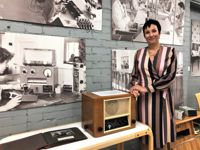 Satu Helkama är nöjd över att man fått till stånd ett fabriksmuseum och att företagets historia börjar finnas väldokumenterad i flera olika historiker. Bilderna i bakgrunden är från utställningen i radiomuseet i Lahtis i samband med företagets 100-årsjubileum.