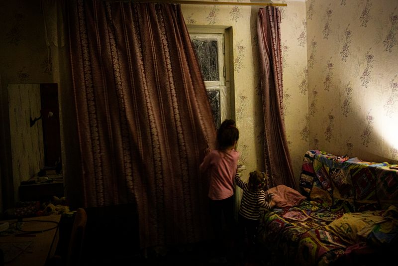 Karolina Bedran, 8, drar för gardinerna i det rum som är sovrum för hela familjen. Fönstren är inplastade för att minska risken för att glassplitter ska flyga in i rummet om en granat slår ner i närheten. Karolina och lillasyster Ariana Ignatenko, 2, kan varken gå i skola eller dagis för tillfället. Det är för farligt att gå i skola eftersom Cherson ständigt beskjuts av ryska trupper på andra sidan Dnjepr. Distansundervisningen fungerar inte heller som den borde eftersom staden är utan el och det inte går att använda surfplattan.