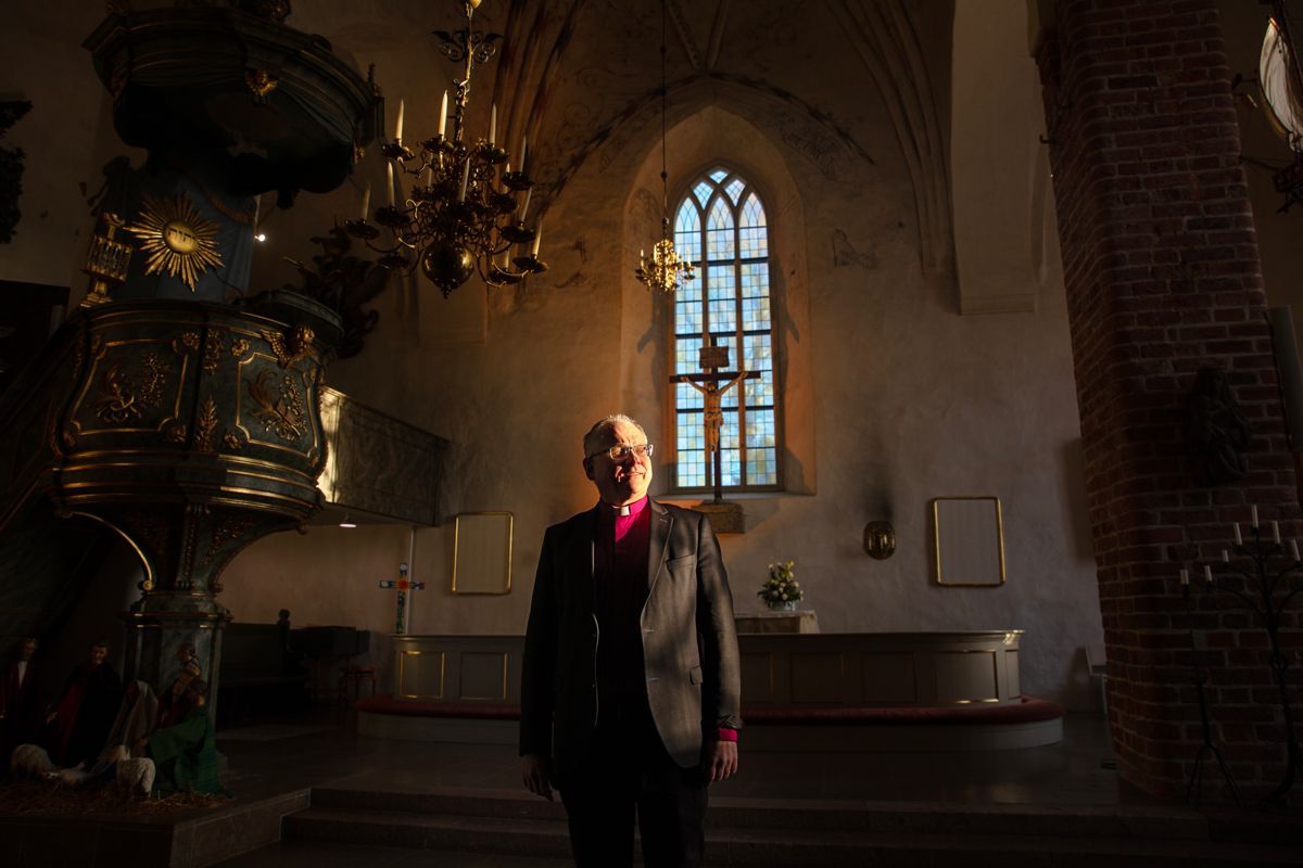Biskopen står i en mörk kyrka, lite solljus kommer in genom fönstrat och lyser på hans ansikte.