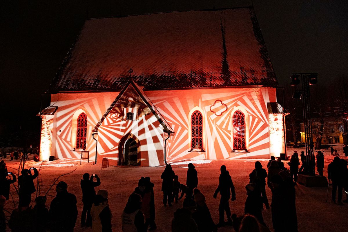 Människor samlade kring kyrkan vars rappade fasad är upplyst med mönster i rött och vitt i kvällsmörkret.