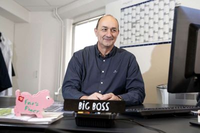 På skrivebordet er der et »navneskilt« hvor der bare står Big Boss. (Allan Ernstrøm er 198 centimeter høj og går lidt i knæ for fotografens skyld) Men selv er slagteri-direktøren meget mere glad for de to malerier, som han har fået af sine medarbejdere ved forskellige anledninger, og som fortæller både om slagteriets og hans personlige historie. Foto: Tom Laursen