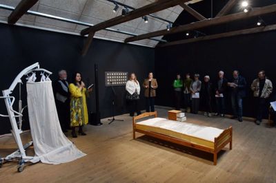En seng, et natbord og en personlift. Kunstpakhusets leder Zana Jankovska (i gult) introducerer udstillingen, mens kunstnerne Maren Kjelgaard og Anna Walther ser til fra hovedgærdet. Foto: Kunstpakhuset