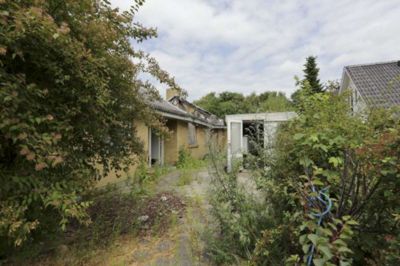 Peter Jessen. Sådan tager brandtomten sig ud set fra vejen. Huset er ved at gro til i meterhøjt ukrudt, men dørene til hus og udhus står åbne