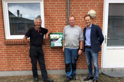 Hjertestarteren hænger nu på østsiden af Rimmerhus Forsamlingshus. Det er Henning Andersen (t.v.), Rimmerhus Maskinforretning, og Morten Mortensen (t.h.), Uni El i Rimmerhus, der har finansieret hjertestarteren, som Richard Andreasen (i midten), Barde El Service, har sørget for ophængningen af. ?Privatfoto