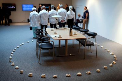 Who cooked Adam Smith dinner? Spørgsmålet blev stillet på ryggen af fem af kunstpavillonens frivillige, der her poserer ved Lise Skous kunstværk af samme navn. Et mødebord, seks stole, en PH-lampe, seks kopper, en kaffekande, en blok og en blyant omgivet af små keramikstykker med håndaftryk fra nogle af bankens ansatte og kunder samt andre Videbæk-borgere. Men det er meget mere end det. Ved ferniseringen fik vi nemlig at vide, at »I sin praksis italesætter Lise Skou sociale og psykologiske konsekvenser ved vores hegemoniske kapitalistiske samfundsstruktur«. Op mod 100 gæster deltog i lørdagens fernisering. ?Foto: Flemming Hansen