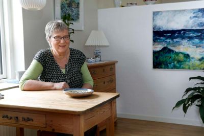 Karin A. Vestergaard sidder ved et bord, der stod i hendes barndomshjem, og hvor hun som lille spiste morgenmad. Maleriet bagved er malet af en ven og tidligere præst. Foto: Henrik Ole Jensen