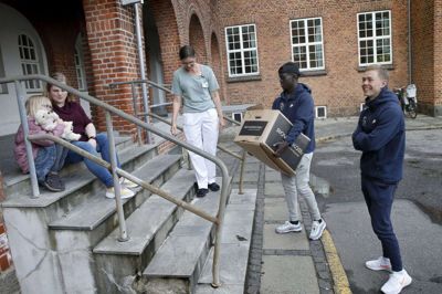 Awer Mabil bærer en kasse fra Legeheltene op til syvårige Victoria, der sidder på trappen med sin mor, Pia Ellesen. Sygeplejerske Mette Linneman tager i mod på vegne af personalet. Joel Andersson satser på, at holdkammeraten selv kan bære kassen. Foto: Flemming Hansen