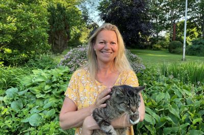 Jette Munkgaard hjemme i sin have med katten Piv på 18 år. Det bedste ved sommeren er de lune aftener herhjemme. I år holder hun ferie i Danmark - i Ebeltoft og skal også på ø-hop til Fur, Endelave og Ærø. Foto: Privatfoto