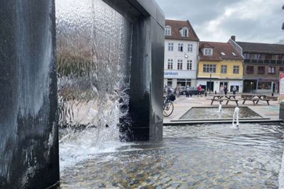 Billedet her er taget fra Torvet og ud mod Storegade. Det er den gule bygning, der er dømt til nedrivning. Der er tale om lidt af et skønbillede med gang i vandkunsten. Den rindende vandkunst er netop sparet bort. Foto: Ikast-Brande Kommune