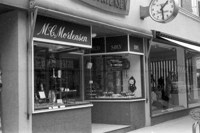 M.C. Mortensens butik fra 1961 med indgang i siden på Missionshotellets ejendom i Bredgade 20. Vest for urmageren lå frisør Heine Sørensen, og til højre anes V. Dalgaards udstilling ud mod hjørnet til Skolegade. Alle butikkerne i hotellets stueplan havde det samme mosaikbeklædte indgangsparti. Bemærk også det tidstypiske cykelstativ på fortovskanten.