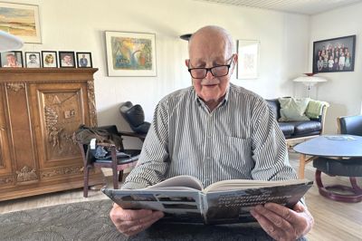 Helge Weis Jensen med første bind af fire i den historiske Vinderup-serie, han udgav i samarbejde med sin nu afdøde hustru, Ninna, der på dækketøjsskabet bag ham våger over familien. Hun var hovedkilden til det materiale, der nu ligger til grund for de tekster, der gemmer sig bag QR-koder på en række historiske bygninger i byen.