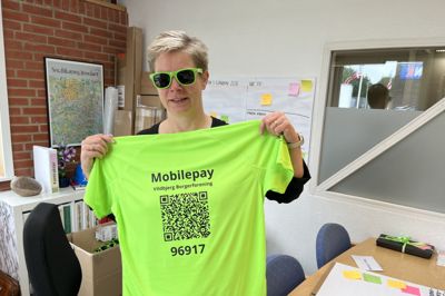 Bykoordinator Birthe Luckmann viser en af de grønne frivilligtrøjer frem. Den smarte QR-kode på ryggen viste sig dog at være lidt for godt kamufleret på den grønne baggrund til, at mobiltelefonerne kunne aflæse den.