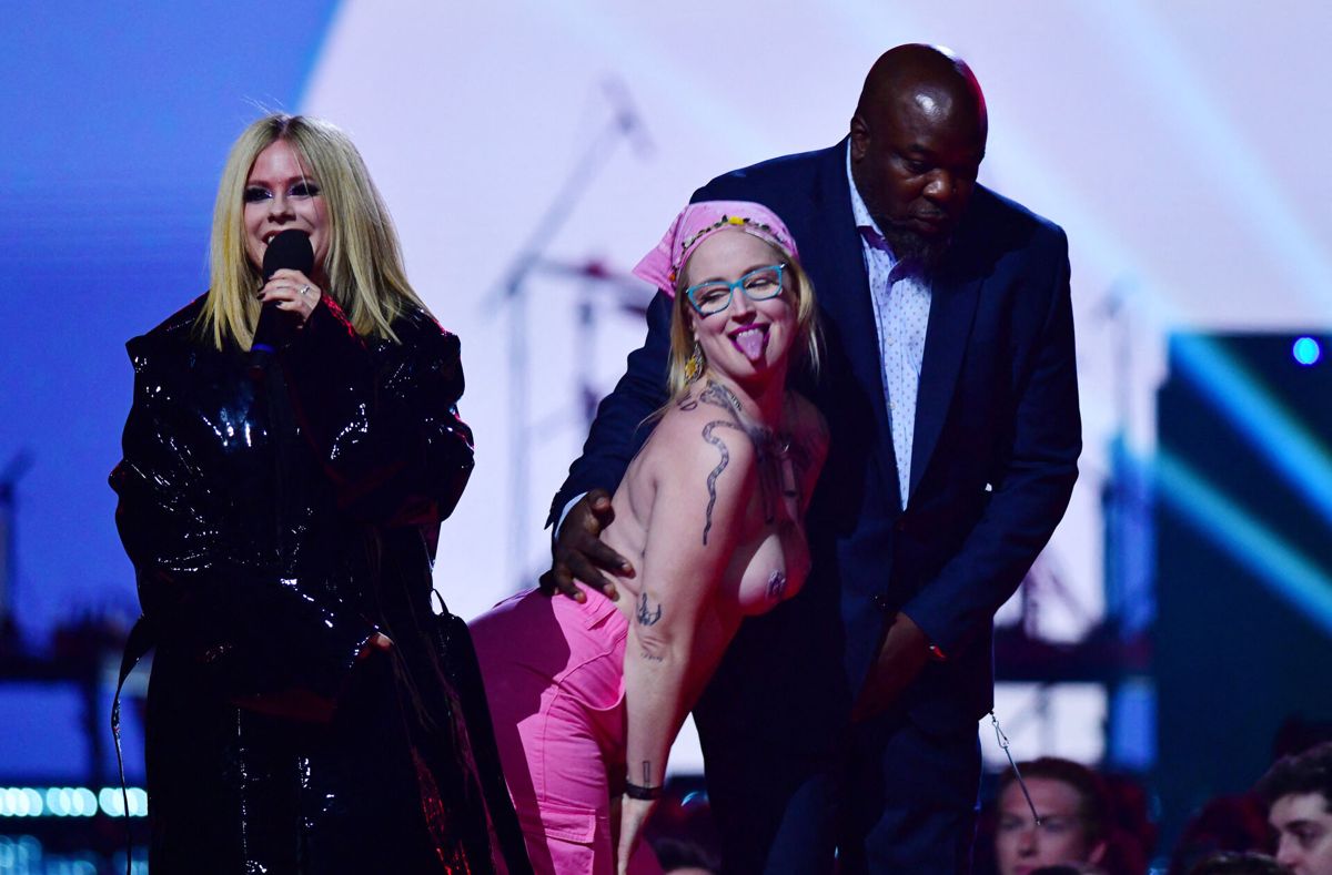 Den topløse miljøaktivist ledsages væk fra scenen af en hærdebred vagt alt imens Avril Lavigne ser smågrinende til.