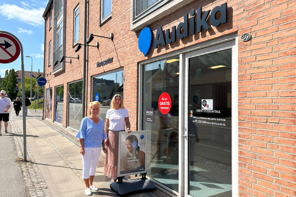 Selvom Dansk HøreCenter Holbæk nu bliver til Audika, møder man som kunde de samme, kendte ansigter, nemlig hørespecialist Lis og kundekoordinator Ulla, som her viser den nye Audika-facade frem.