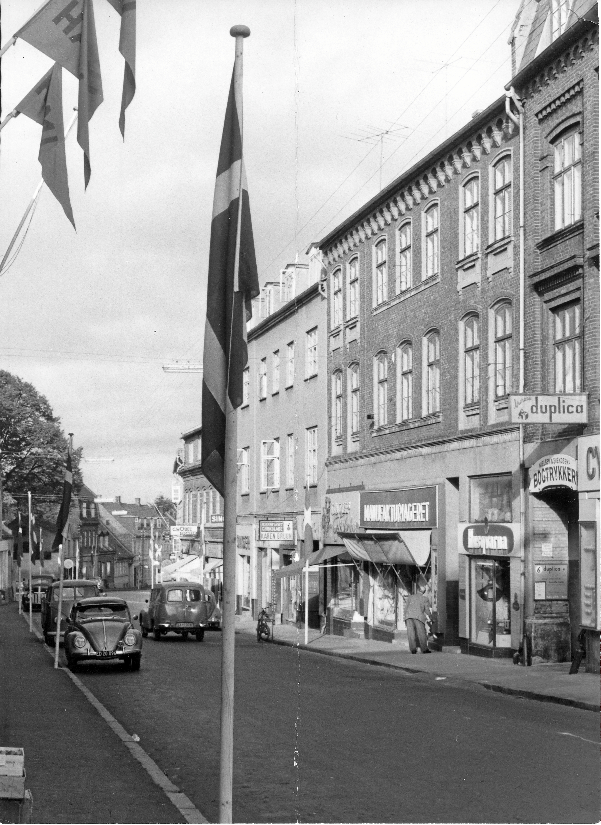 Løvegade 8, hvor Ruth og Erik Gøtze begyndte i 1945. På facaden ses skilte for: Cyclecompagniet, Bogtrykkeri Nielsen & Svendsen, Duplica, Manufakturlageret, Gøtzes fiskehus og Karen Bruuns konfekture. - Arkivleder Christoph Klinger, Slagelsearkiverne forklarer, at billedet er fra årene 1958-1965, da nummerpladen på bilem tidligst kan være anvendt i 1958, bogtrykkeriet ikke medtaget i vejviser 1965/66.